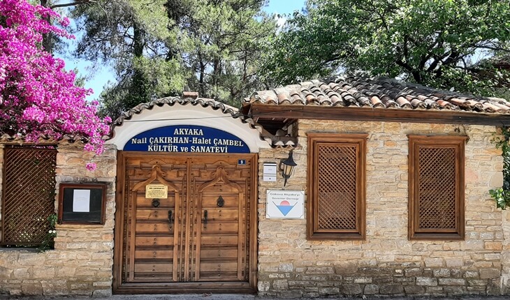 Akyaka’daki taş duvarlı, ahşap kapılı Nail Çakırhan-Halet Çambel Kültür ve Sanat Evi’nin dış cephesi; yanında begonvil çiçekleri ve çam ağaçları görülüyor.